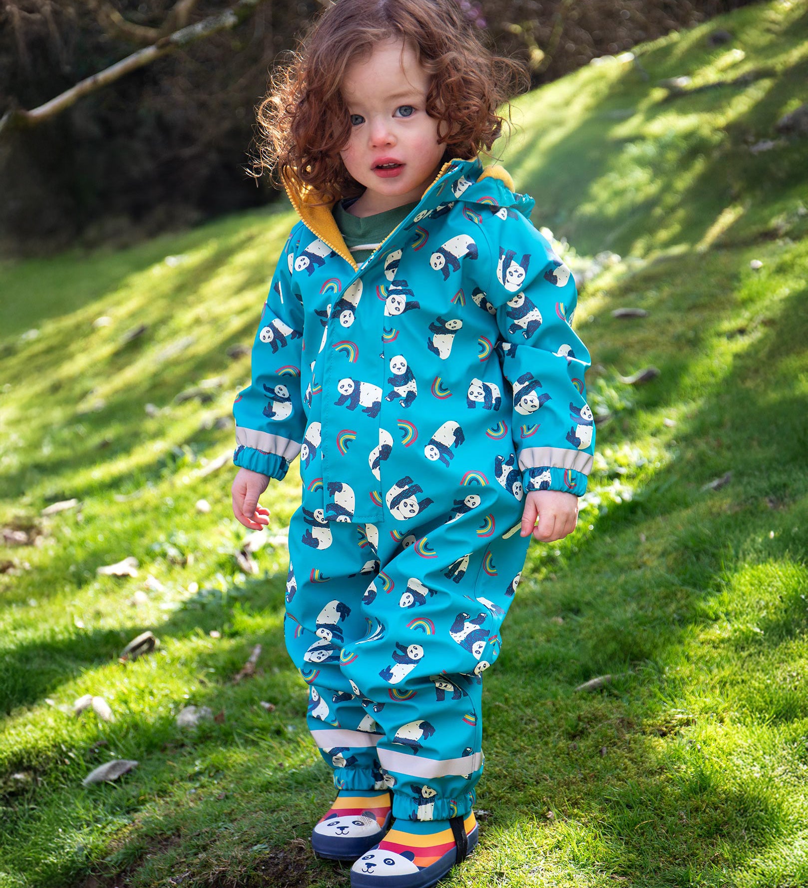 A toddler wearing the Frugi puddle buster blue panda play waterproof all in one suit with the panda wellingtons.