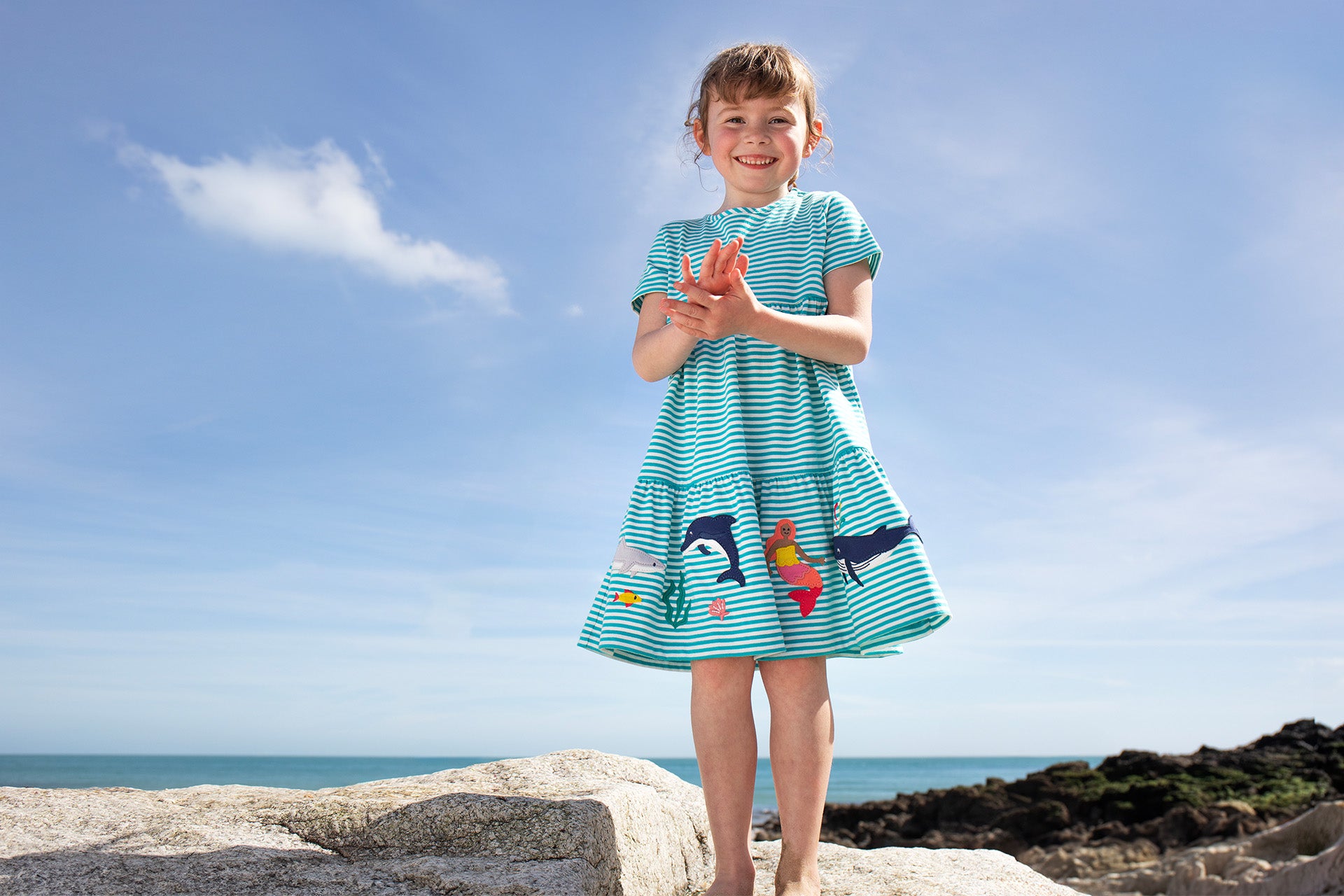 Girl on beach wearing a Frugi Summer dress with dolphins and mermaids. Organic cotton summer clothing at babipur