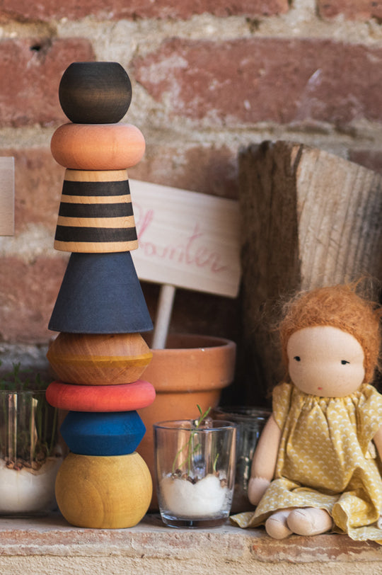 Stack of colorful wooden blocks by Grapat Toys with a doll sitting next to them against a brick wall.