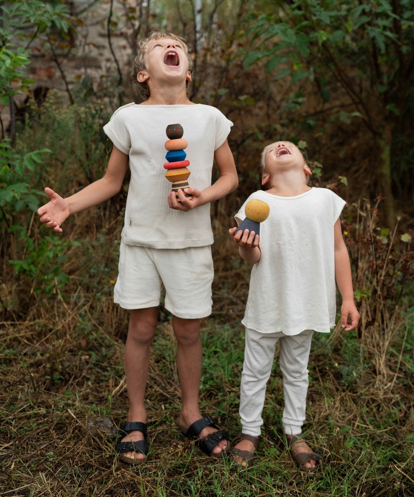 2 children standing on a grassy floor in the garden holding the pieces from the Grapat wooden serendipity stacking set. Features a brown, light pink, blue yellow orange and striped pieces.