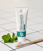 The Green People Fresh Mint & Aloe Vera Toothpaste with Fluoride is stood on a white tiled floor, with a bamboo tooth brush in front and a sprig of fresh mint leaves to the side
