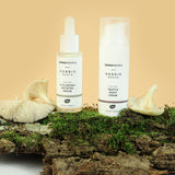 The Green People Nordic Roots Hyaluronic Booster Serum and Truffle Night Cream are placed upright on a wooden log with moss and chanterelle mushrooms, to represent the natural ingredients used in the Nordic Roots range