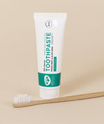 The Green People Natural, Fresh Mint & Aloe Vera Toothpaste with Fluoride on a beige background with a bamboo toothbrush in front of the toothpaste tube