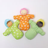 Three Grimm's Waldorf Baby doll toys laid out next to each other. The left doll Baby Leo has a light green body with white flower print, a light blue hat, and light brown hair. The middle doll Baby Mia, has an orange body with white flower print, a light pink hat and white hair, and the left doll Baby Sam has a green check print body, a yellow hat and black hair.