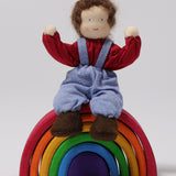 A closer look at Peter, sat on a Grimm's small wooden rainbow, with its arms bent upwards to show the dolls flexibility, on a light grey background. Peter is a handmade, Waldorf, fabric doll.