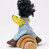 A view of Grimm's Handmade Doll - Max from the back. Max is climbing over a Grimm's small natural rainbow to show how flexible its arms and legs are. The image is on a light grey background