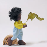 Grimm's Handmade Doll - Max is stood next to a Grimm's small natural rainbow and is holding a green leaf in its hand. The image is on a grey background