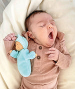 A small baby is yawning whilst sleeping, and is cuddles with Grimm's Lavender Doll Aqua