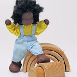 Grimm's Handmade Doll - Max is standing next to a Grimm's small natural rainbow, with its foot on one of the pieces so show how Max can move its arms and legs. The image is on a light grey background