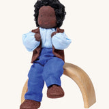 Grimm's Handmade Doll - Mr Ebony, is sitting on a natural rainbow arch. The doll has black skin, black curly hair, a smiling face, and is wearing brown fabric shoes, blue trousers, a blue top and a brown sleeveless jacket. The image is on a cream background.