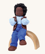 Grimm's Handmade Doll - Mr Ebony, is sitting on a natural rainbow arch. The doll has black skin, black curly hair, a smiling face, and is wearing brown fabric shoes, blue trousers, a blue top and a brown sleeveless jacket. The image is on a cream background.