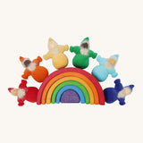 The Grimm's Rainbow Dwarfs toy dolls sat on top of a grimm's small wooden rainbow toy, on a cream background
