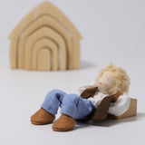 The Grimm's Blond Haired Man Doll, laying down on its back with its arms behind its head, and its knees bent in a relaxed position. There is a Grimm's Natural House in the background.