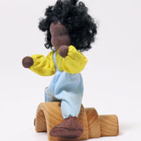 A side view of Grimm's Handmade Doll - Max sitting on a Grimm's small natural rainbow. The image is on a light grey background