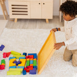 A child is sat on a fluffy cream rug, and has tipped the blocks of the Grimm's Basic Building Set from their wooden base