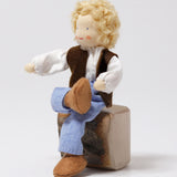 The Grimm's Blond Haired Man Doll is sat down on a block of wood with its legs crossed, showing the flexibility of the doll