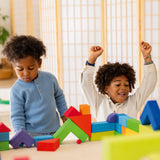 Two children are playing with the Grimm's Basic Building Set. One child has their hands up in the hair and looks excited for what they have built, and the other child is concentrating on pushing a small blue marble under a block shaped like a bridge
