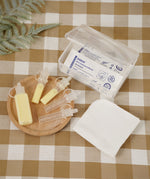 The Haakaa Silicone Colostrum Collector multi-size pack is sat on a  small light brown wooden dish, with a small open box with Silicone Colostrum Collectors inside in their packaging. Underneath is a table cloth in cream and brown check print