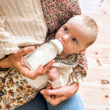 Baby feeding from a Hevea glass baby bottle on parents lap.