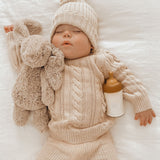 Baby wearing a cream knitted hat and jumper asleep with a Hevea Glass Baby Bottle and toy rabbit.