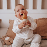 Baby sat in a cot smiling with the Hevea Glass Baby Bottle Wide Neck With Rubber Sleeve - 150ml in Sand.