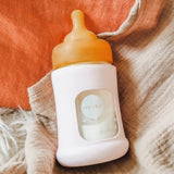 Hevea Glass Baby Bottle Wide Neck With Rubber Sleeve - 150ml in Sand on cream and terracotta muslins.