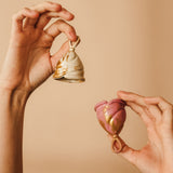 Person holding up two menstrual cups, one cream and gold, the other pink and gold.