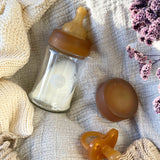 A Hevea Glass Baby Bottle half filled with milk, a natural rubber cap and dummy on a white muslin.