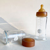 Hevea Wide Neck Glass Baby Bottle Natural Rubber Cap next to a glass baby bottle.