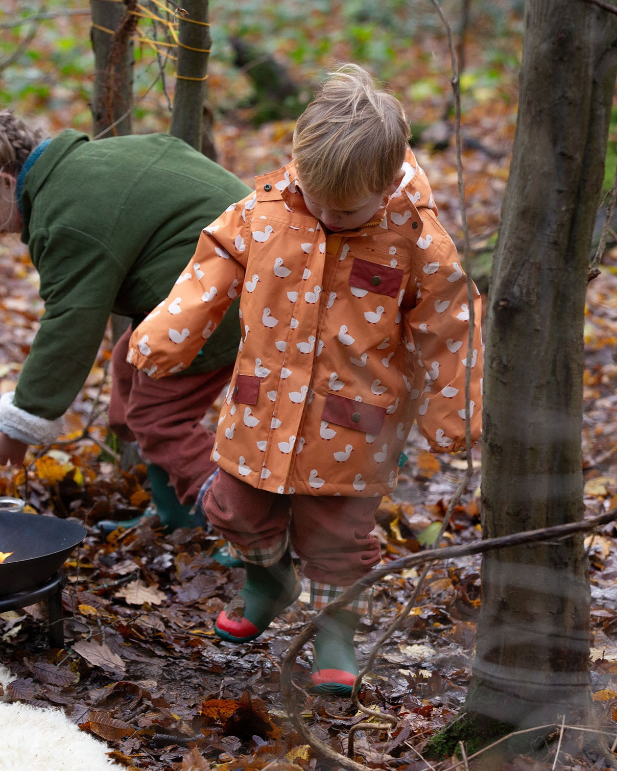Child at a campfire in a forest wearing a Kids Warm Waterproof Winter School Coat in a gold yellow duck print, blue and floral, by Little Green Radicals, made from recycled bottles and a cosy polyester fleece, buy now at Babipur.