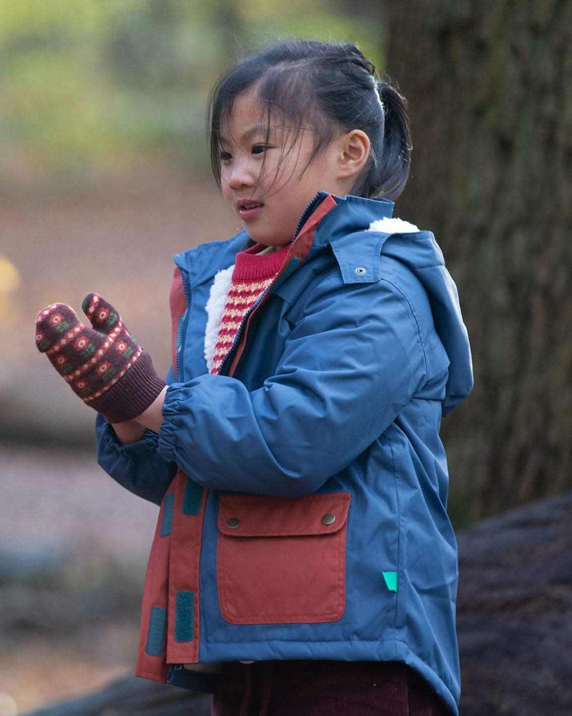 Child in a forest wearing a Kids Warm Waterproof Winter School Coat in a Moonlight Blue print with deep red details down the zip and on the front pockets, PFC Free, by Little Green Radicals, made from recycled bottles and a cosy polyester fleece, buy now at Babipur.
