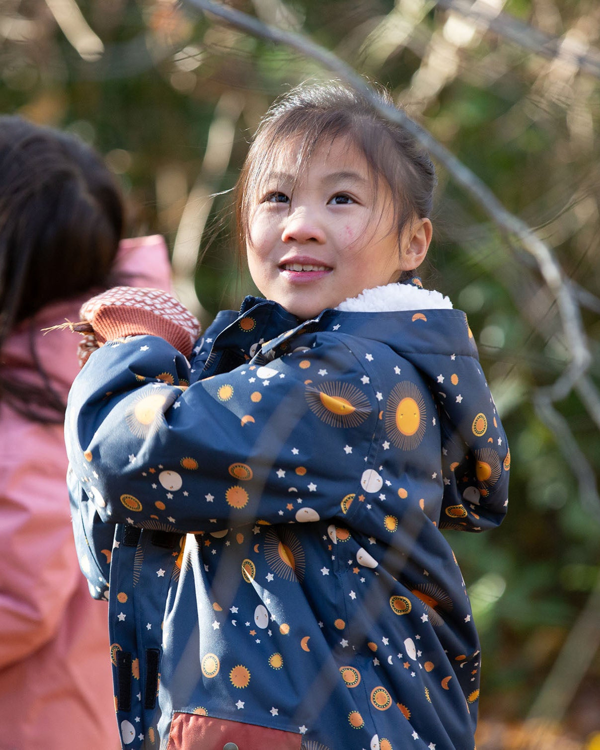 Child in a forest wearing a Little Green Radicals Waterproof Winter Coat with a cosy fleece hood, made from recycled bottles and a cosy polyester fleece, in a navy Night Moon print, available now at Babipur.