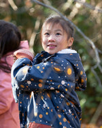 Child in a forest wearing a Little Green Radicals Waterproof Winter Coat with a cosy fleece hood, made from recycled bottles and a cosy polyester fleece, in a navy Night Moon print, available now at Babipur.