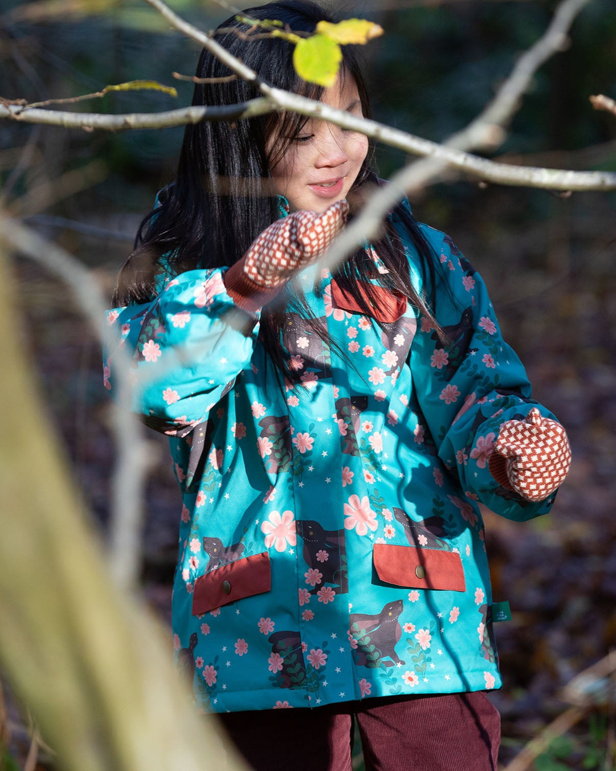 Girl stood in a forest wearing a Warm and Waterproof (5000M rating), PVC free kids school coat by Little Green Radicals, in a Winter Rabbit blue print, with red front pockets, buy now at Babipur.