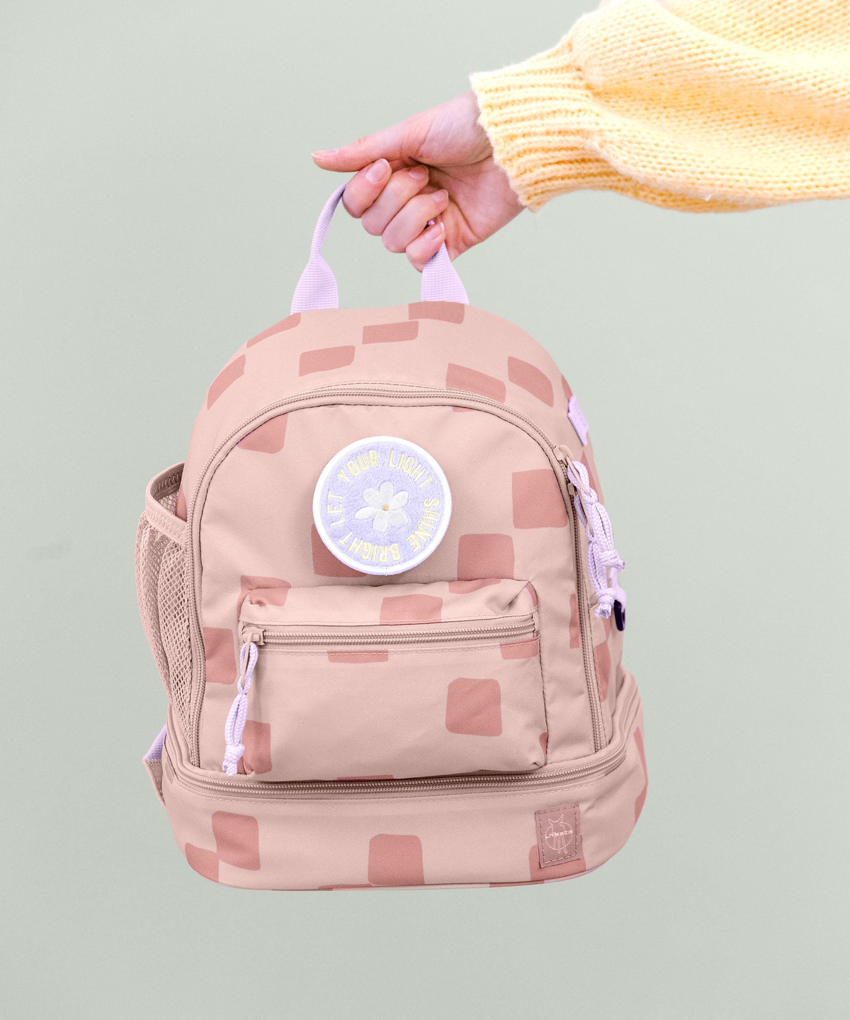 A adult hand holding the Lassig kids mini pattern party rose pattern backpack by the lilac handle on the top of the backpack