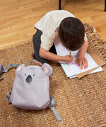 A child sitting on a rug, drawing next to the Lassig koala tiny backpack.
