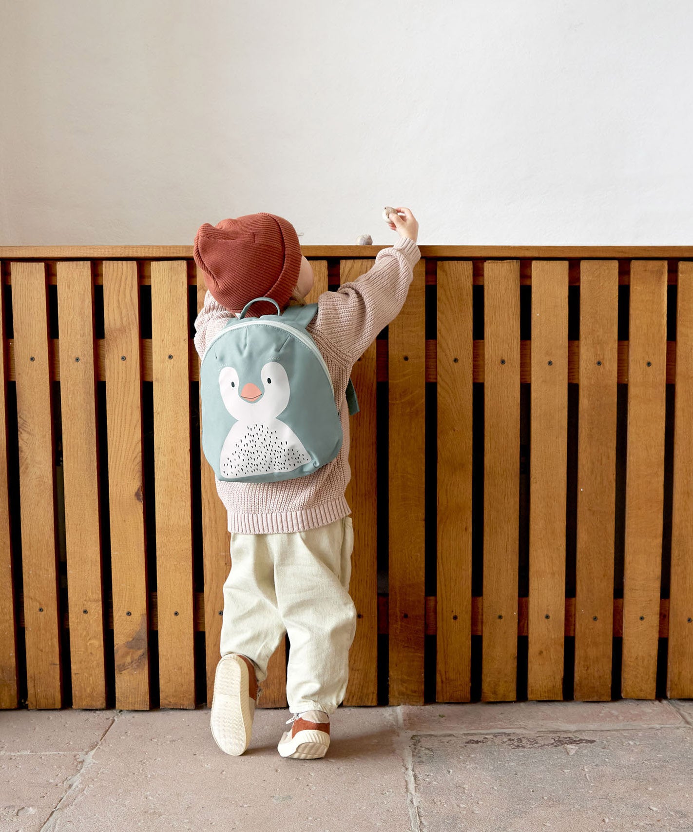 A small child standing near a wooden fence and wearing the Lassig kids tiny backpack in penguin