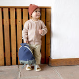 A small child standing near a wooden fence and holding the Lassig kids tiny backpack in whale by the top handle