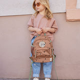 A child standing outside and holding the Lassig kids mini caramel happy pattern backpack by the handy loop handle on the top of the backpack