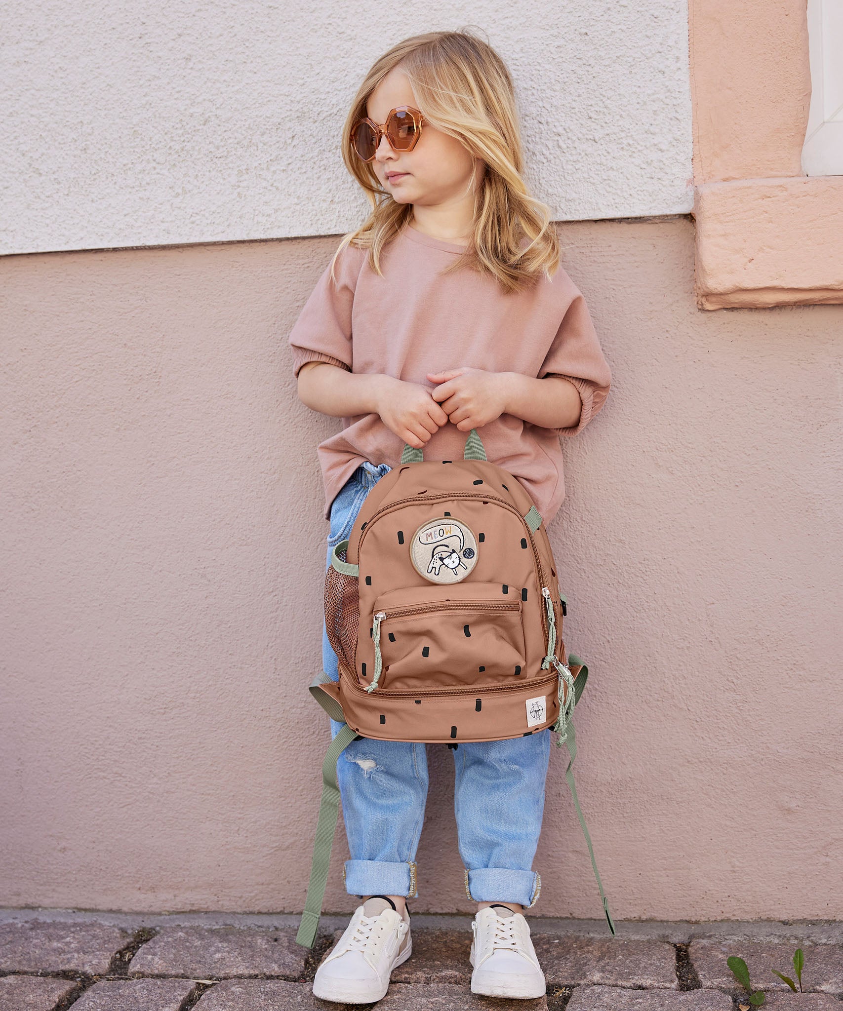 A child standing outside and holding the Lassig kids mini caramel happy pattern backpack by the handy loop handle on the top of the backpack