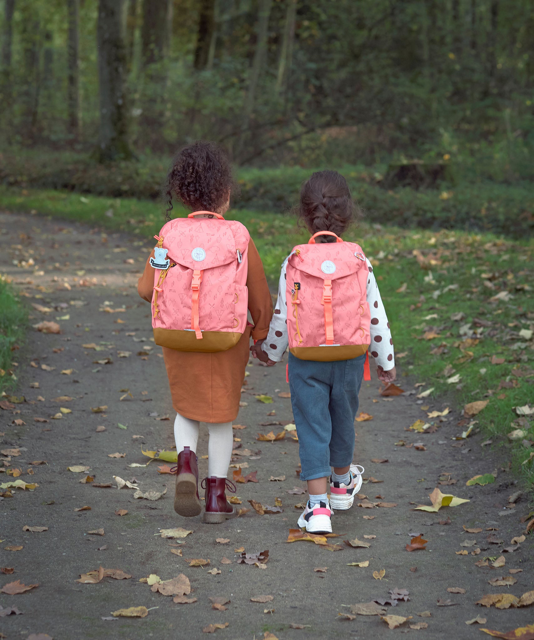 Two kids walking through a forest and wearing the Lassig kids mini adventure backpack in rose
