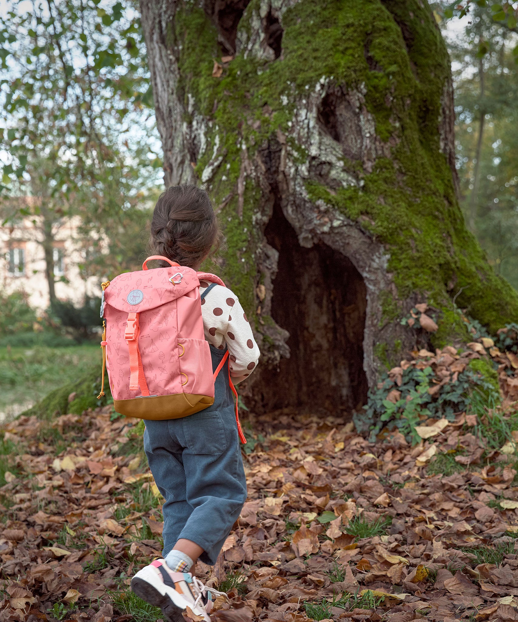 A child walking through leaves near a big tree and wearing the Lassig kids mini advetnure backpack in rose.