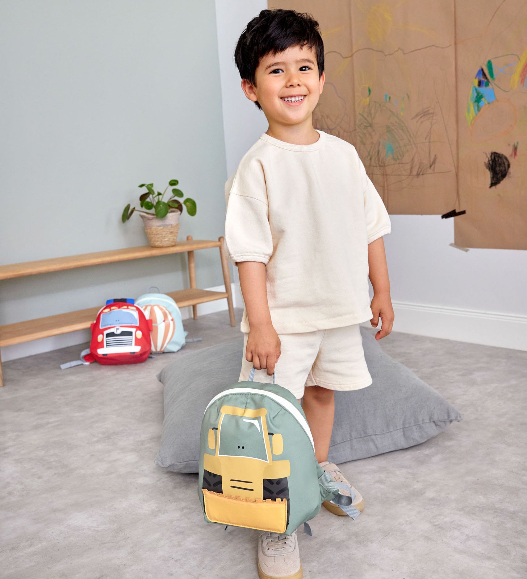A child in a playroom and holding the Lassig kids tiny digger backpack by the loop handle on the top.