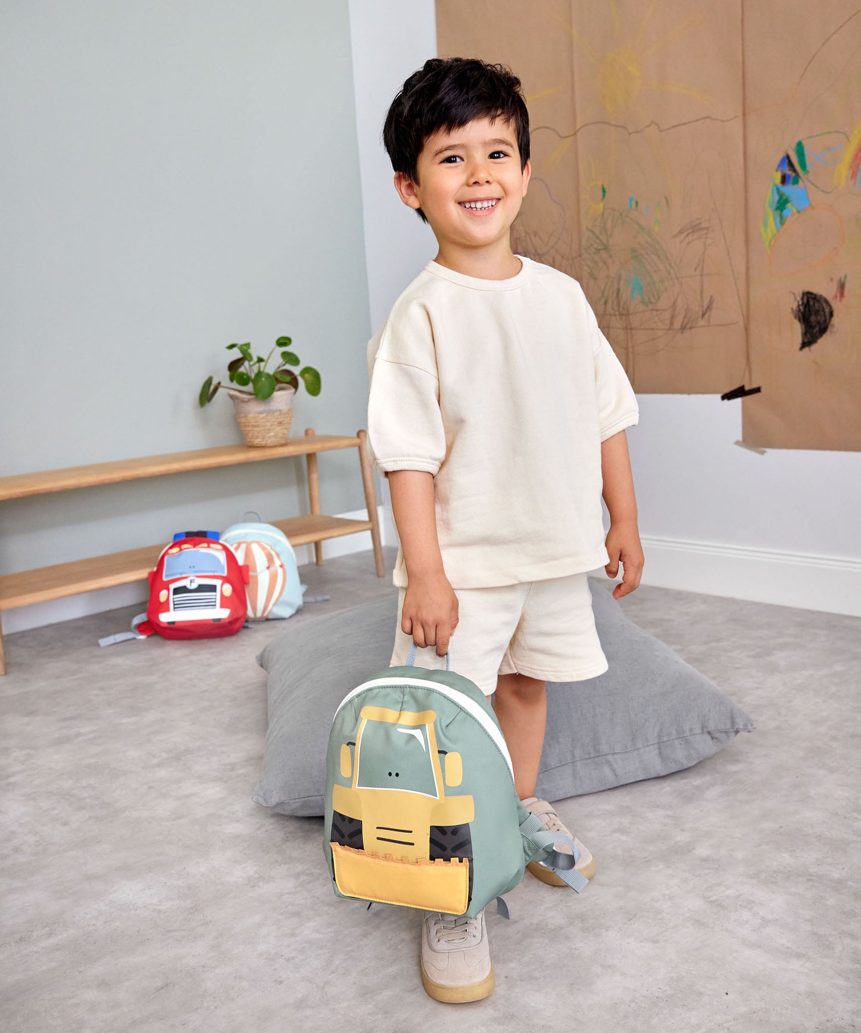 A child in a playroom and holding the Lassig kids tiny digger backpack by the loop handle on the top.