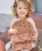 A child sitting outside and holding the Lassig kids mini happy prints Caramel pattern backpack. A pale brown backpack with a cute horse patch on the front panel.