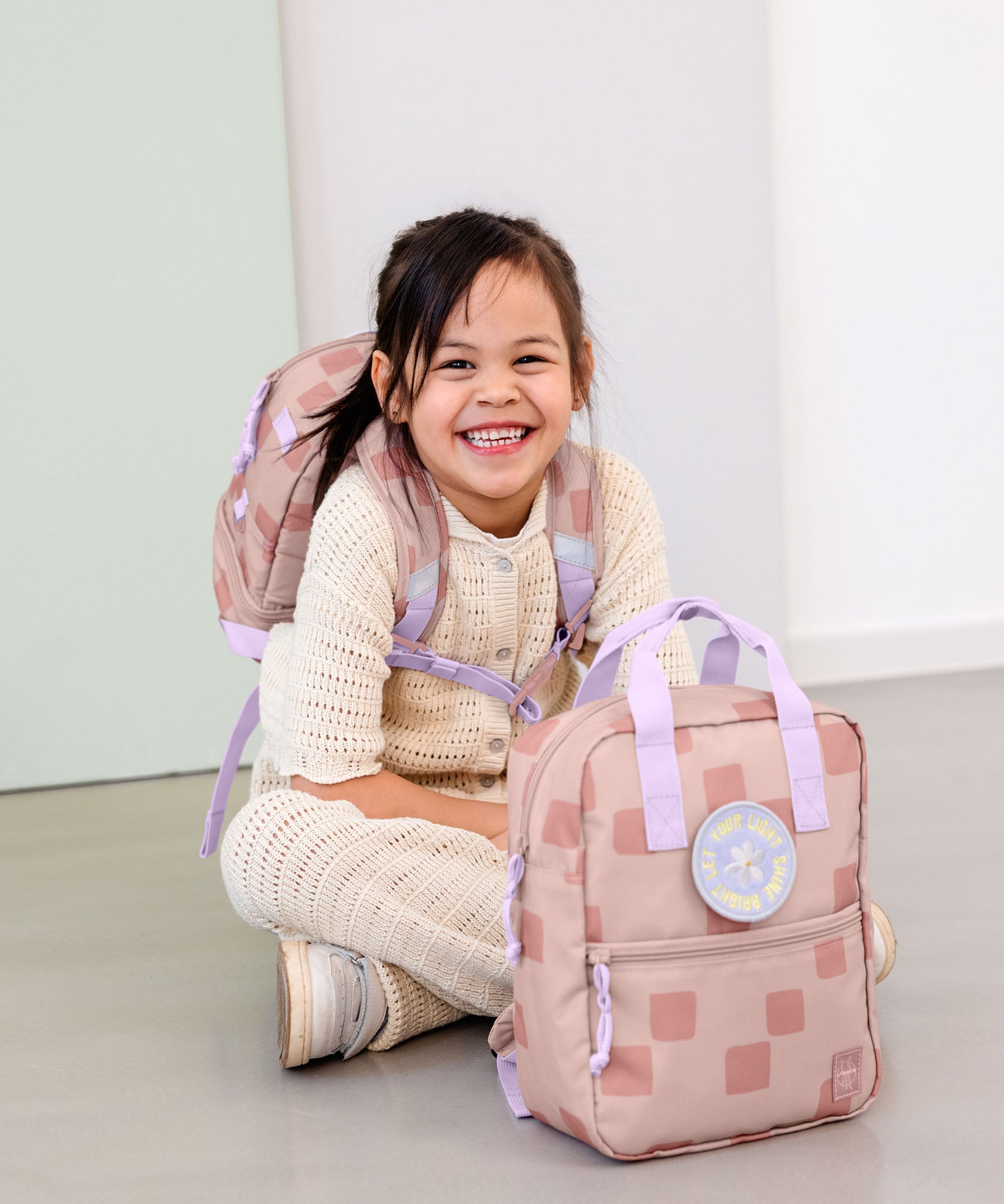 A child sitting on the floor and wearing the Lassig kids mini pattern party rose pattern backpack with another in front.