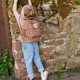 A child standing by a wall and wearing the Lassig kids mini happy prints caramel backpack