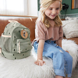 A child sitting on a couch next to the Lassig kids mini happy prints olive backpack