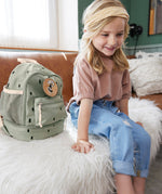 A child sitting on a couch next to the Lassig kids mini happy prints olive backpack