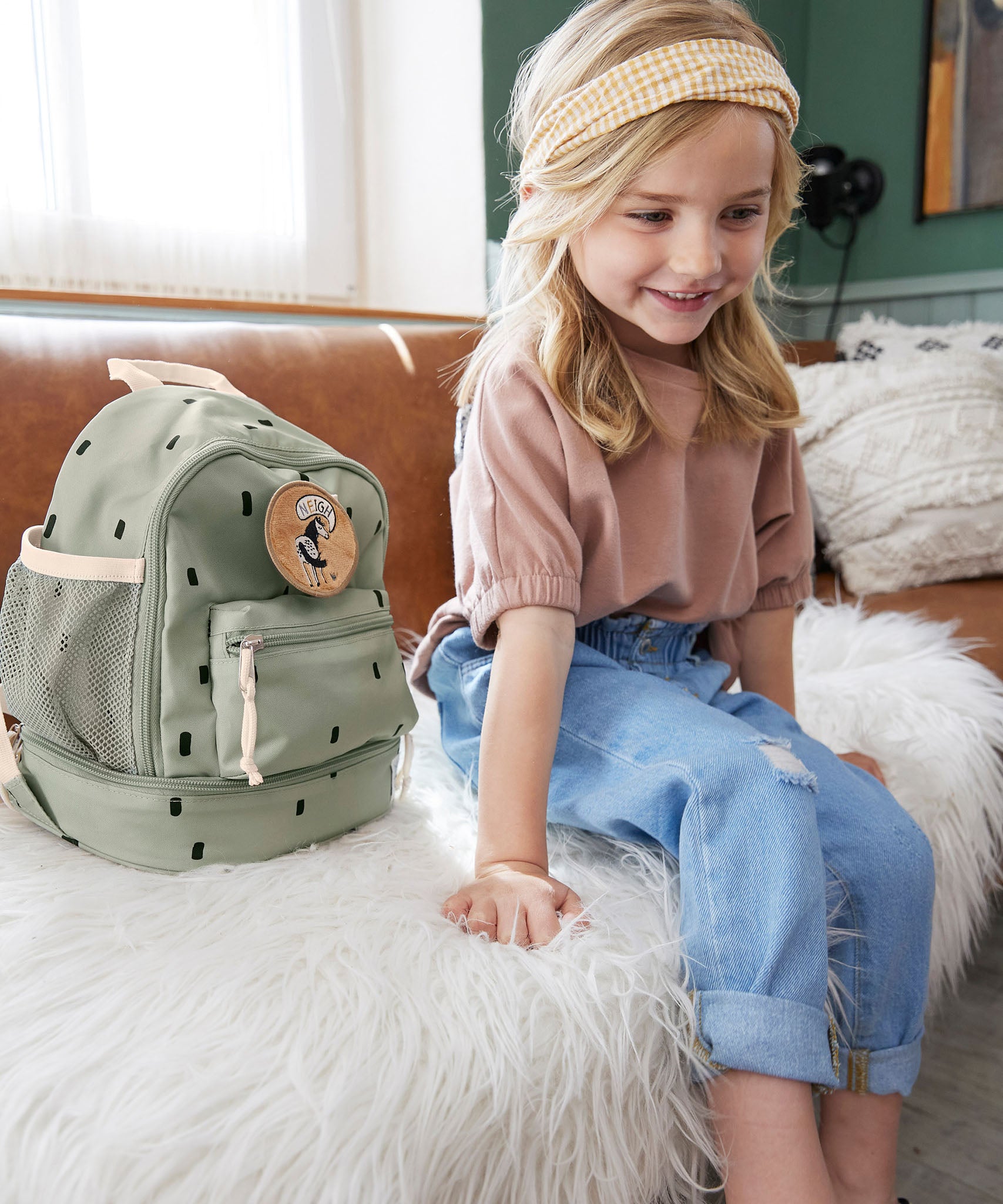 A child sitting on a couch next to the Lassig kids mini happy prints olive backpack